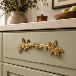 May include: A gold-colored metal drawer pull featuring an ornate, symmetrical leaf-like design with intricate detailing. The handle is mounted horizontally on a flat, light sage green cabinet drawer front with a smooth, matte finish.