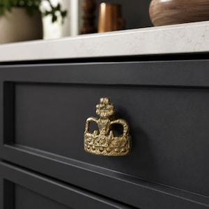 May include: A gold-colored metal cabinet knob shaped like a crown with a textured, antique finish. The crown features intricate detailing along the base and top, mounted onto a dark charcoal gray drawer front with a flat, matte surface finish.