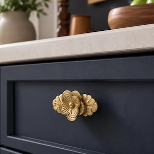 May include: A gold-toned metal cabinet knob shaped like a flower with layered petals and leaves, mounted on a dark navy blue drawer front. The hardware features a textured, matte finish with a central circular detail and defined petal lines.