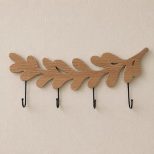 May include: A wall-mounted coat rack featuring a horizontal wooden piece shaped like a leafy branch with a natural light brown wood grain finish. Four thin, black metal hooks are attached to the bottom edge of the wooden branch for hanging items.