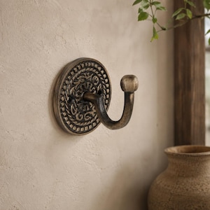 May include: A wall-mounted metal hook featuring a circular base plate with an intricate, embossed pattern and a curved arm ending in a rounded knob. The finish is a dark, weathered bronze tone with lighter highlights on the raised details.