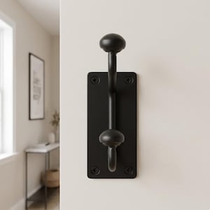 May include: A black metal coat hook mounted on a rectangular plate. The hook has a rounded top and a curved hook at the bottom. The plate is attached to the wall with four screws. The hook is designed for hanging coats, hats, or other items.