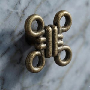 May include: A brass-toned metal cabinet knob featuring a symmetrical, knotted design with four circular loops extending from a central vertical bar. The hardware has a weathered, antique finish and is mounted onto a grey and white marble surface.