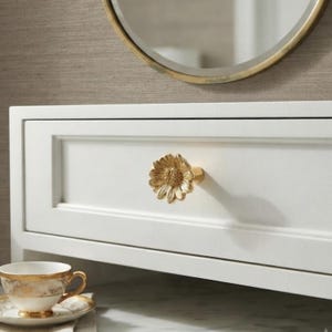 May include: A gold-toned metal drawer knob shaped like a flower with detailed petals and a textured center, mounted on a white rectangular drawer front. The knob features a metallic finish and a dimensional design that protrudes from the flat surface.