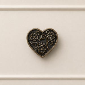May include: A heart-shaped cabinet knob with a floral design. The knob is made of dark metal with an antique finish. The flowers and vines are intricately detailed, adding a touch of elegance to any furniture piece.