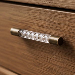 May include: A drawer pull featuring a clear, textured, cylindrical glass center with a twisted, diamond-cut pattern. The handle is finished with two cylindrical, antique brass-toned metal end caps, mounted horizontally on a light brown wood surface.