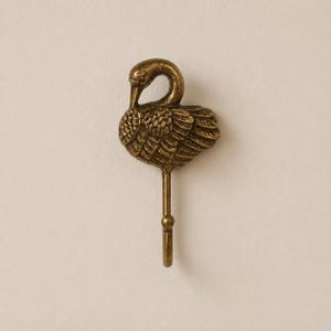 May include: A brass-colored swan-shaped wall hook. The hook features a detailed swan with its head tucked down, and a small hook at the bottom. The hook is designed to be mounted on a wall for hanging items.