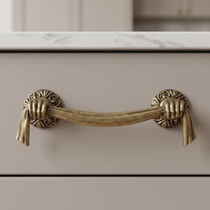 May include: Antique brass drawer pull with a unique design. The pull features a curved handle held by two decorative circular plates, each adorned with a hand holding a draped fabric. The hardware is mounted on a light gray cabinet.