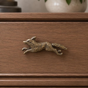May include: A brass-toned metal drawer pull shaped like a running fox with a textured, weathered finish. The fox is depicted in a horizontal, leaping pose with its head turned slightly forward, mounted onto a flat, medium-brown wooden surface.