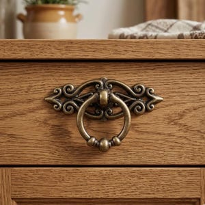 May include: Close-up of a bronze-colored drawer pull with a ring handle and ornate scrollwork design. The hardware is mounted on a wooden drawer, showcasing the details of the furniture's construction.