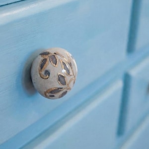 May include: A round ceramic drawer knob featuring a cream-colored base with a hand-painted pattern of grey leaves outlined in metallic gold. The knob is attached to a light blue painted wooden drawer front with a smooth, matte finish.
