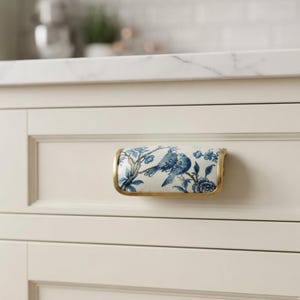 May include: A rectangular drawer pull featuring a white ceramic surface with a blue botanical pattern of birds perched on branches. The ceramic insert is framed by a metallic gold-toned border and is mounted on a cream-colored cabinet drawer.