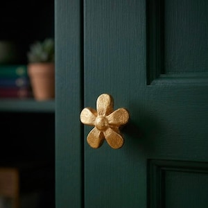 May include: A gold-colored metal cabinet knob shaped like a five-petaled flower with a textured, matte finish. The knob is mounted on a dark forest green wooden cabinet door with a visible vertical grain and a recessed panel design.