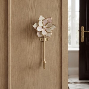 May include: A wall-mounted hook featuring a decorative flower-shaped top with layered, translucent pale pink and white petals and a central pearl-like accent. The hook has a slender, brushed gold-tone metal stem and a circular mounting plate.