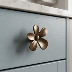 May include: A flower-shaped cabinet knob with a metallic bronze finish and a textured surface. The knob features five rounded petals radiating from a central circular point, mounted on a light blue wooden drawer front with visible wood grain.