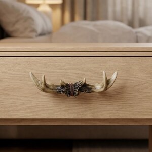 May include: A drawer pull shaped like a pair of light brown deer antlers with a dark brown textured center band, mounted horizontally on a light-colored wooden drawer front with a visible horizontal wood grain pattern.
