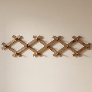 May include: Wooden accordion-style wall-mounted coat rack. The rack is made of light brown wood and features a diamond-shaped design. Small, round, gold-colored knobs are at each intersection. The rack is mounted on a neutral-colored wall.