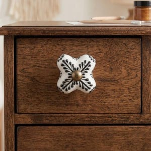 May include: A white ceramic drawer knob with a four-lobed shape and a black floral pattern. The center of the knob features a small, circular, metallic bronze-toned accent piece. The knob is mounted on a dark brown wooden drawer front.