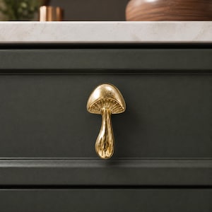 May include: A gold-colored mushroom-shaped cabinet knob with a smooth, reflective finish and visible gill details under the cap. The knob is mounted on a dark green drawer front, featuring a curved stem and a rounded cap design for furniture.