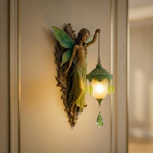 May include: A wall-mounted light fixture featuring a bronze-toned fairy figure with green wings. The fairy holds a lantern with a frosted glass shade, green leaf-shaped accents, and a dangling green crystal teardrop pendant that emits a warm glow.