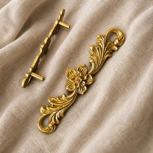 May include: Two gold-colored metal cabinet pulls featuring an ornate floral and leaf design. The primary pull is displayed horizontally, showcasing a central flower with detailed petals and scrolling foliage extending to each end with mounting posts.