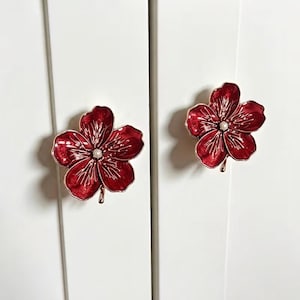 May include: Two flower-shaped cabinet knobs with a deep red enamel finish and metallic gold-toned edges. Each knob features five rounded petals with embossed vein details and a small white pearl-like bead centered in the middle of the flower.