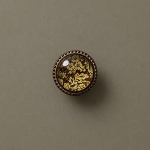 May include: A round cabinet knob with a floral design. The knob features a clear glass center with a yellow floral pattern on a dark brown background. The knob is surrounded by a bronze-colored metal rim with small, decorative beads.