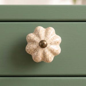 May include: A flower-shaped cabinet knob with a cream-colored, crackled glaze finish and a central metallic bronze-toned screw head. The knob is mounted on a flat, solid sage green surface, providing a clear contrast between the textured ceramic.