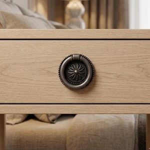 May include: A light oak wood drawer front featuring a circular, dark metallic drop pull handle. The handle has a beaded outer edge and a central floral motif with twelve petals. The wood grain is horizontal and visible across the drawer surface.