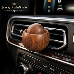 May include: A wooden car air vent diffuser with a matching wooden sphere inside. The diffuser and sphere are made of a dark brown wood. The car's interior features a black dashboard with silver accents. The text "JewelryDisplayStudio" is at the top.