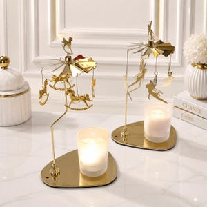 May include: Two gold-coloured metal candle spinners with a reflective base. One spinner features hanging rocking horse ornaments, while the other features hanging fairy ornaments. Each unit includes a clear glass votive candle holder with a lit candle.
