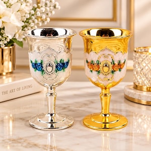 May include: Two ornate metal goblets, one with a silver finish and one with a gold finish. Each goblet features a white enamel band decorated with raised, colourful floral motifs and a central oval emblem, set on a slender stem and base.