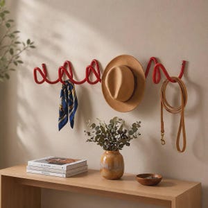 May include: A red metal wall-mounted coat rack with a continuous, winding, spiral-like design. The rack holds a tan fedora hat, a blue and gold patterned silk scarf, and a tan leather leash. The structure features multiple loops for hanging items.