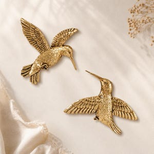 May include: Two gold-colored hummingbird wall decorations with detailed feather textures. One bird is positioned with wings angled upward, while the other bird is depicted with wings spread horizontally. Both figures feature pointed beaks.