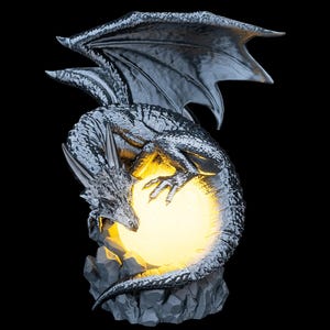 May include: A silver-colored dragon figurine perched on a rocky base, featuring textured scales and large wings. The dragon is curled around a glowing, spherical light source that emits a warm yellow illumination, highlighting its detailed form.