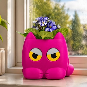 May include: A bright pink planter shaped like a stylised cat with large, yellow, expressive eyes and pointed ears. The planter holds a small potted plant with green leaves and clusters of white and purple flowers, sitting upright on a flat surface.