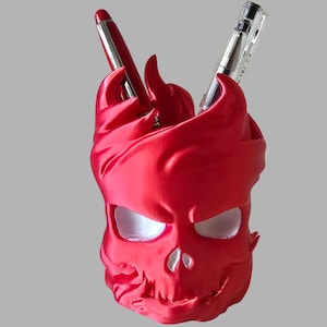 Könnte beinhalten: Ein leuchtend roter Stiftehalter in Totenkopfform mit flammenartigem Design am oberen Rand. Die hohlen Augen- und Nasenhöhlen sind weiß. Zwei Stifte stecken in der oberen Öffnung, einer mit rotem Gehäuse und einer mit transparentem Gehäuse.
