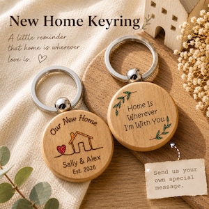 May include: Two round wooden keychains with silver metal rings. One features a house outline, a red heart, and the text "Our New Home Sally & Alex Est. 2026". The other displays the text "Home Is Wherever I'm With You" with green leaf accents.