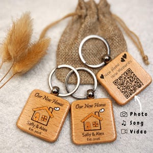 May include: Wooden keychains with silver key rings. The keychains have a square shape with a house graphic and the text "Our New Home" and "Sally & Alex Est. 2026". One keychain has a QR code and the text "New Home New Memories".