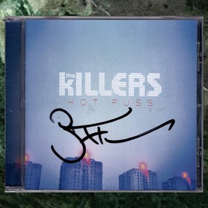 May include: A CD case featuring the album "Hot Fuss" by The Killers. The cover art displays the band's name in white pixelated letters above the album title in red. The case is signed in black marker. The background shows a cityscape.