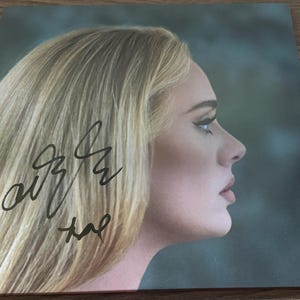 Op de afbeelding: Een vierkante vinyl platenhoes met een profielportret van een blond persoon met lang haar. Een zwarte inkt handtekening staat aan de linkerkant onderaan de afbeelding. De achtergrond is een donkere, gedempte kleur blauwgroen met zachte belichting.