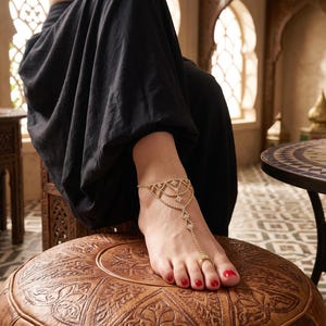 Könnte beinhalten: Fußkette in Gold mit Zehenring und dekorativen Elementen. Der Fuß mit rotem Nagellack ruht auf einem braunen Lederhocker. Die Person trägt ein schwarzes Kleidungsstück. Orientalisches Thema.