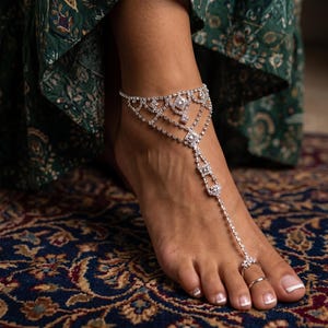 Könnte beinhalten: Eine silberne Fußkette mit Zehenring und mehreren rautenförmigen Verzierungen. Der Schmuck wird an einem Fuß mit weißer Pediküre getragen. Der Fuß ruht auf einem gemusterten Teppich.