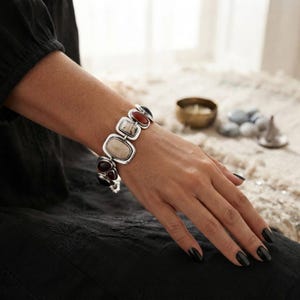 May include: A silver-toned link bracelet featuring a series of rectangular and oval-shaped stones in shades of cream, deep red, and black. The stones are set within polished metal frames, creating a continuous chain design that wraps around the wrist.
