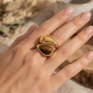 Può includere: Un anello bypass color oro caratterizzato da due elementi a forma di goccia. Un elemento è in metallo color oro liscio e lucido, mentre l'altro è una pietra preziosa occhio di tigre marrone e oro con un distinto motivo a strisce cangianti.