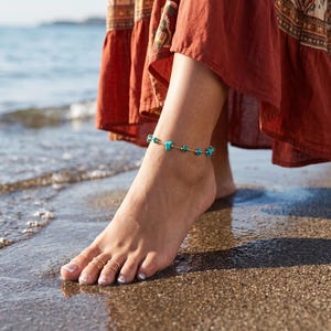 Può includere: Braccialetto alla caviglia con perline turchesi e dettagli color oro. Il gioiello è indossato su un piede sulla spiaggia. La persona indossa un lungo vestito color ruggine. L'oceano è sullo sfondo.