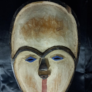 Op de afbeelding: Een uit hout gesneden masker met een lichtgekleurd gezicht en een gladde, bleke afwerking. Het masker heeft amandelvormige oogopeningen die felblauw zijn geschilderd, een donkere driehoekige neus en een verticale rode streep vanaf de neus.