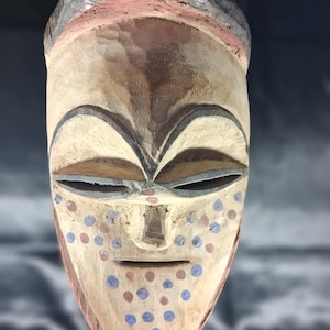 Op de afbeelding: Een handgesneden houten masker met een glad, lichtgekleurd gezicht, donkere, gebogen wenkbrauwen en amandelvormige ogen. Het onderste deel van het gezicht is versierd met een patroon van kleine, ronde blauwe en bruine stippen en een donkerbruine kroon.