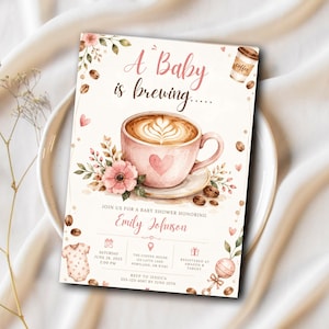 May include: A rectangular baby shower invitation card featuring a watercolor illustration of a pink coffee cup with latte art, surrounded by coffee beans, pink flowers, a baby onesie, and a rattle. The text reads A Baby is brewing, Emily Johnson, and event details.