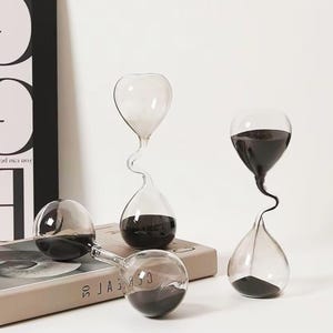 May include: Four clear glass hourglasses with black sand inside. The hourglasses have a unique, curved design. One hourglass is lying on its side, while the others are upright. The background is a neutral color.